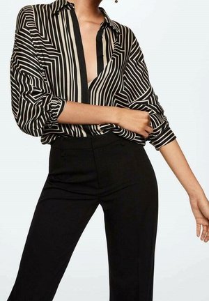 Personne portant une chemise rayée verticale noire et blanche à manches bouffantes et un pantalon noir ajusté, posant avec un bras plié.