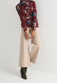 Maglione floreale bordeaux con collo a cowl, abbinato a pantaloni larghi crema e stivaletti alla caviglia verde oliva. Tessuti morbidi e motivi vivaci.