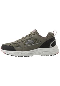 Zapatilla deportiva verde oliva con parte superior de malla, acentos negros y entresuela blanca. Punta y lazo en el talón texturizados para facilitar el uso.