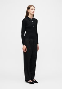 Γυναίκα που φοράει μαύρο μακρυμάνικο πουκάμισο με γιακά και κουμπιά, φαρδιά μαύρη παντελόνα και μαύρα μυτερά φλατ παπούτσια, στέκεται μπροστά σε απλό λευκό φόντο.