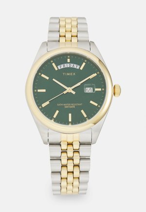 Timex wristwatch with green dial, gold and silver metal band, day display showing Friday, date showing 14, and gold hour markers and hands.