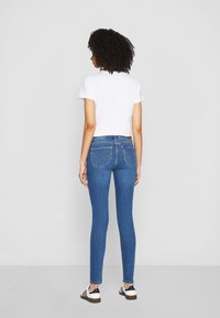 Jeans in denim blu slim fit con vita media, caratterizzati da due tasche posteriori e un motivo di cucitura sottile, abbinati a una maglietta bianca a maniche corte.