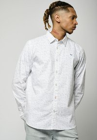 Chemise blanche à manches longues avec un petit motif floral bleu, boutonnière à l'avant et un logo discret sur la poitrine. Tissu doux avec un col classique.