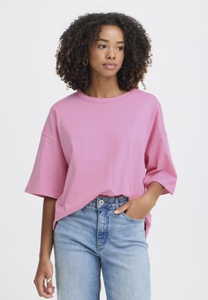 Kobieta o kręconych włosach, ubrana w luźną różową koszulę z krótkim rękawem włożoną do jasnoniebieskich, wysokich dżinsów, stojąca z jedną ręką w kieszeni.