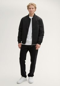 Giacca bomber nera con polsini e colletto a costine, indossata su una maglietta bianca, abbinata a jeans neri e sneakers bianche. Design minimalista.