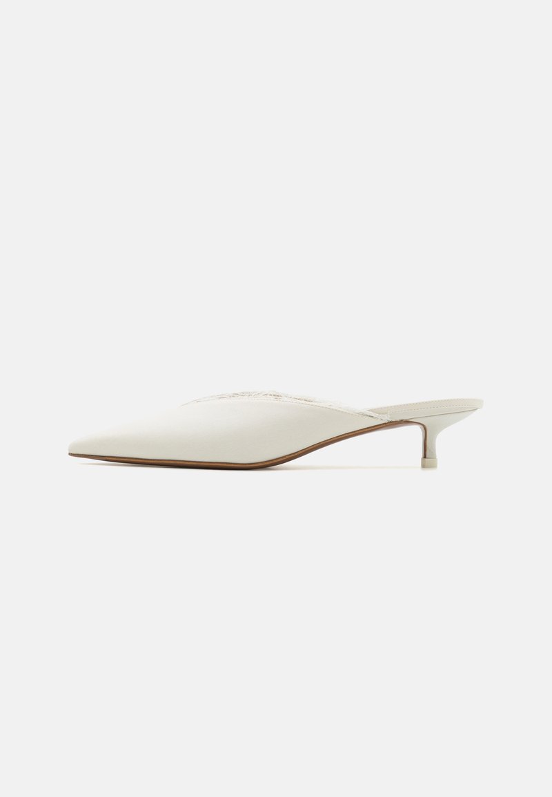 Mule de punta afilada en tejido blanco roto. Presenta un tacón bajo y un sutil detalle de costura a lo largo del empeine. Textura suave, diseño minimalista.