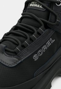 Tenisi negri cu glezna înaltă, cu un finisaj texturat, având șireturi proeminente, un logo și accente gri contrastante pe materialul superior.