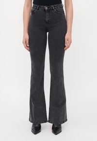 Kvinne iført svarte flagrende jeans og svarte spisse støvler, står mot en hvit bakgrunn.