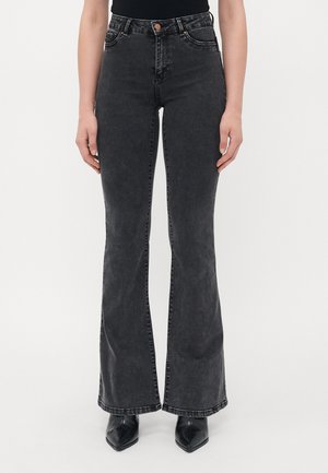 Kvinne iført svarte flagrende jeans og svarte spisse støvler, står mot en hvit bakgrunn.