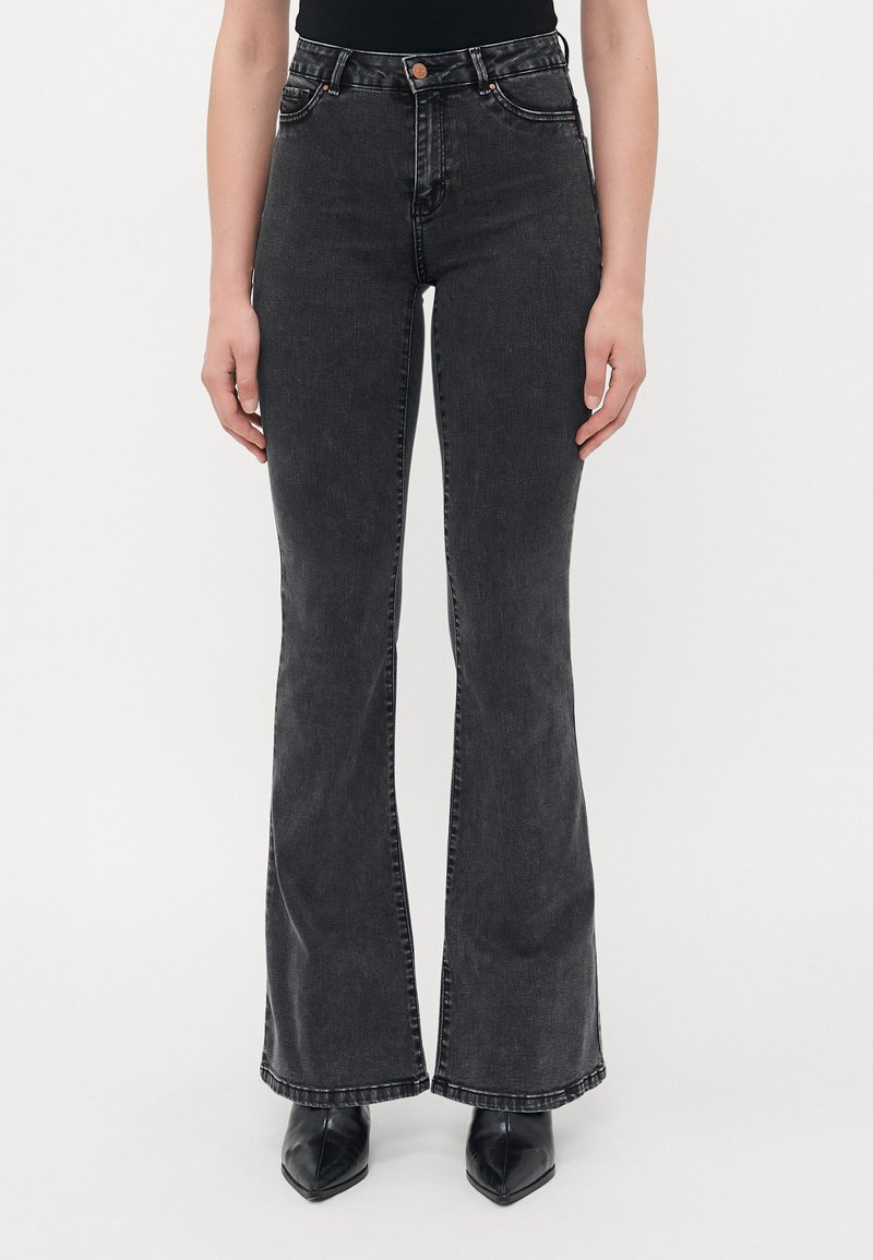 Kvinne iført svarte flagrende jeans og svarte spisse støvler, står mot en hvit bakgrunn.