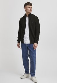 Jachetă neagră cu fermoar, cu textură ribbed orizontală, purtată peste un tricou alb, asortată cu blugi albaștri și adidași albi.
