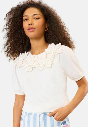 Woman with voluminous curly hair wearing white blouse with 3D flower details and blue-and-white striped skirt with floral accents.