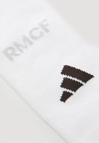 Biele plátno s textúrovaným nápisom "RMGC" v svetlosivej farbe a tmavohnedým trojuholníkovým logom so bielymi akcentmi na spodnej časti.