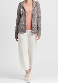 Felpa grigia con texture e cordini crema, indossata su una t-shirt rosa, abbinata a pantaloni bianchi a gamba larga e scarpe sportive beige.