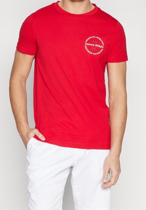 Mann trägt ein rotes Tommy Hilfiger T-Shirt mit weißer Schrift auf der Brust und weiße Hose, Hand in der Tasche.