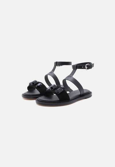 Μαύρα επίπεδα σανδάλια με λουράκια στον αστράγαλο, γεωμετρικές διακοσμήσεις στα μπροστινά λουράκια και ασημένια πόρπη.