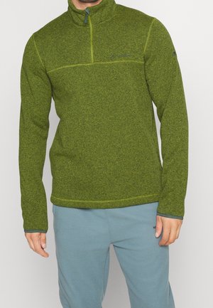 Muž ve svetru z olivově zeleného fleece s čtvrtým zipem a světlých modrých volnočasových kalhotách stojící před jednoduchým pozadím.