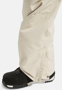 Beige sneeuwbroek met een wijd beenontwerp, verstelbare enkelopening over een zwarte sneeuwboot, voorzien van drukknoopsluiting en een gladde textuur.