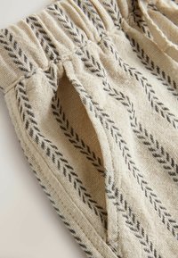Țesătură bej cu model negru în zigzag, având o talie elastică și buzunar lateral, care evidențiază textura și detaliile cusăturilor.