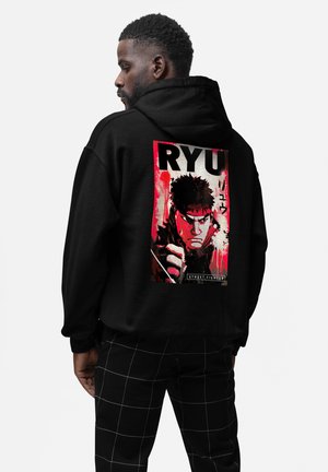 Uomo con capelli corti e ricci che indossa una felpa con cappuccio nera con una grafica rossa e nera di Ryu di Street Fighter sul retro, abbinata a pantaloni a quadri neri.