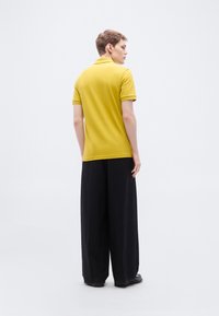 Polo jaune à manches courtes, associé à un pantalon noir ample, vu de dos avec une texture lisse et mate.