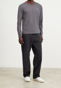 Pitkähiha, harmaa puuvillapaita, jossa pyöreä pääntie ja logo. Yhdistetty löysiin mustiin housuihin ja valkoisiin lenkkikengiin. Minimalistinen muotoilu.