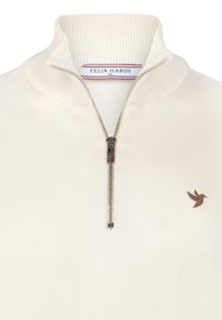 Sweater crème zippé avec un col montant, présentant une texture côtelée, une fermeture éclair en métal et un petit logo d'oiseau brun sur le côté gauche.