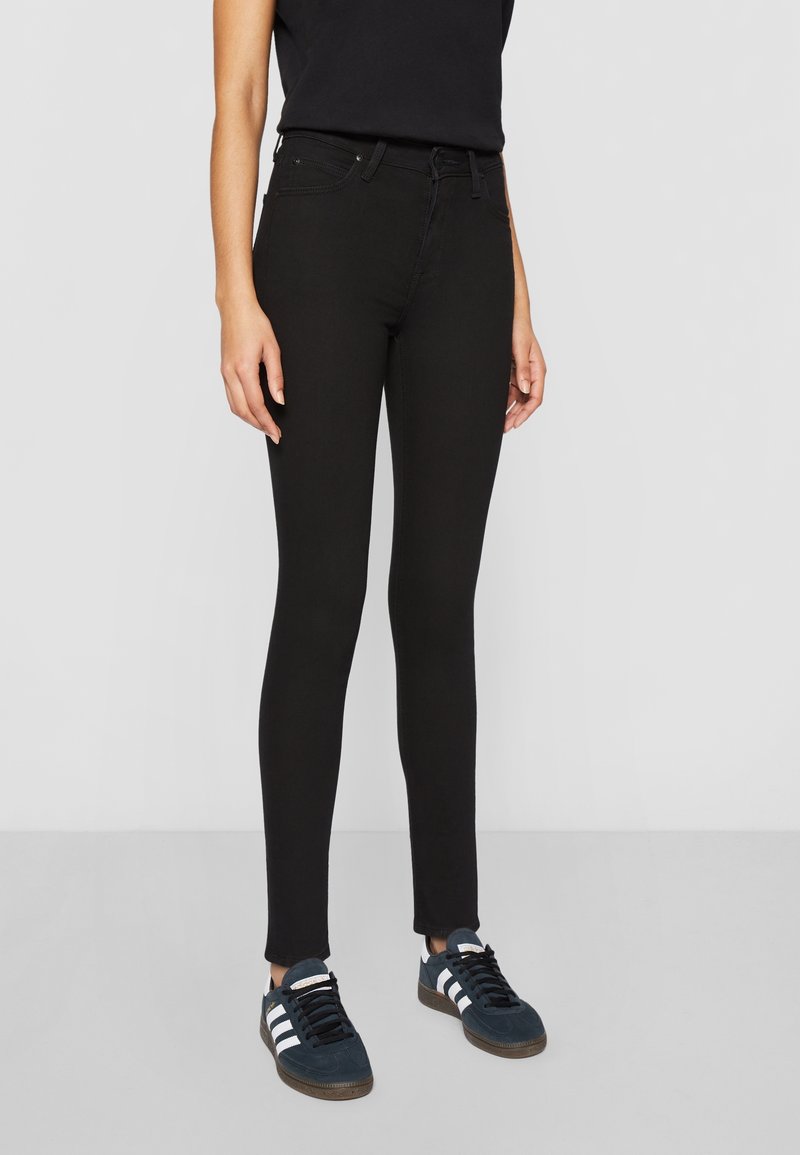 Jean skinny noir avec une texture lisse, taille haute et coupe slim, associés à des baskets sombres avec des rayures blanches.