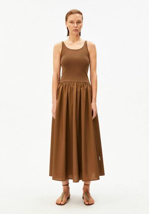 Woman standing facing forward wearing a sleeveless brown dress with a fitted top and full, gathered skirt, paired with flat sandals.