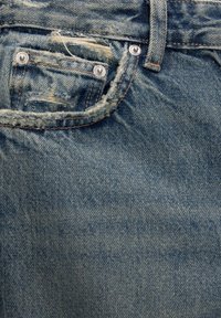 Jeans de mezclilla con un color azul desgastado, textura desgastada y un bolsillo frontal con remaches de metal y bordes deshilachados.