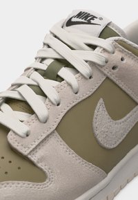 Κοντινό πλάνο σε αθλητικό παπούτσι Nike σε ελιάς πράσινο και μπεζ χρώμα με επενδύσεις από σουέτ, λευκά κορδόνια και λογότυπο Nike στο πλάι και στη γλώσσα.