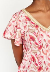 Halenka s květinovým vzorem ve světle růžové a krémové barvě, s krátkými volánovými rukávy a zlatým lemem kolem výstřihu. Měkká tkanina.
