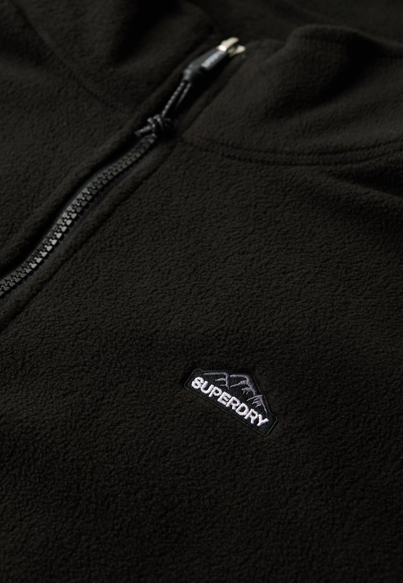 Zwarte fleece zip-up jack met een zachte textuur, voorzien van een Superdry-logo in het wit en grijs nabij de rits. Duurzame rits hardware.