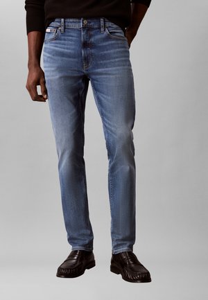 Άνδρας που φορά στενά μπλε τζιν και σκουριά καφέ δερμάτινα παπούτσια χωρίς κορδόνια, στέκεται μπροστά σε ουδέτερο φόντο.