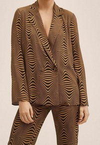 Kobieta w brązowej i czarnej marynarce w geometryczny wzór oraz dopasowanych spodniach, stojąca z rękami luźno wzdłuż ciała.
