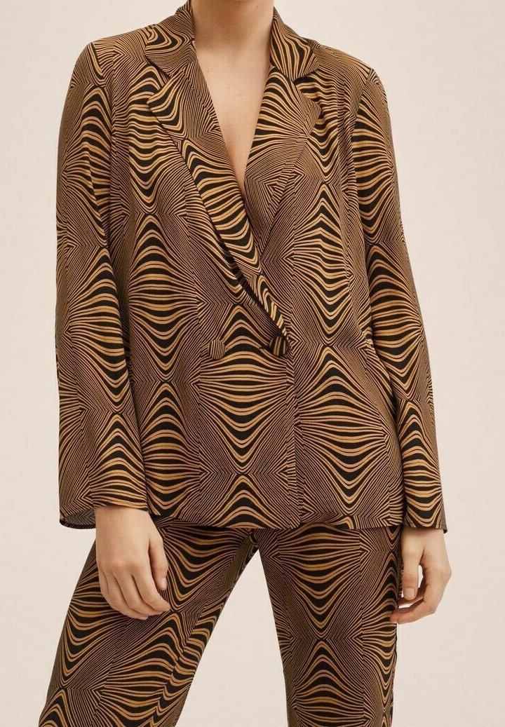 Kobieta w brązowej i czarnej marynarce w geometryczny wzór oraz dopasowanych spodniach, stojąca z rękami luźno wzdłuż ciała.