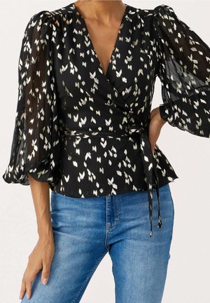 Zwarte wikkelblouse met doorschijnende, wijd uitlopende mouwen en witte bloemblaadjesachtige patronen. Gemaakt met een ceintuur in de taille, gecombineerd met blauwe spijkerbroeken.