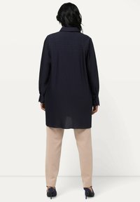 Camicia a pieghe blu navy con colletto, maniche lunghe e polsini con bottone, abbinata a pantaloni beige, che mostrano una texture liscia e un design classico.