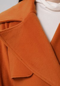 Manteau en laine orange avec un large col et une texture douce, superposé sur un haut à côtes blanc. La vue rapprochée met en avant les détails du tissu.