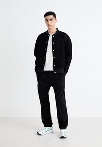 Blouson noir avec des boutons-pression argentés, t-shirt blanc texturé, pantalon noir et chaussures de sport avec des accents verts. Design minimaliste.