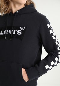 Sweat à capuche noir avec un grand patch logo texturé portant le texte "Levi's". Détails à motif damier sur les deux manches. Poche kangourou.