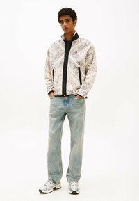 Giacca in pile bianca con motivo, dotata di cerniera nera, abbinata a jeans azzurri chiari e sneaker grigie. La giacca presenta disegni intricati.