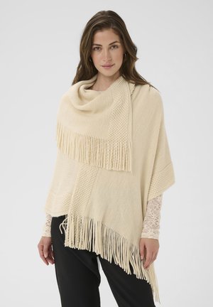 Beige poncho gemaakt van gestructureerde stof met een gedrapeerde hals, met een franjeszoom en een geweven patroon langs de voorkant.