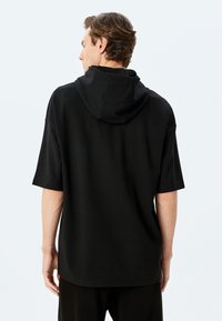 Sudadera negra con capucha, de manga corta, con textura suave, corte relajado y dobladillo redondeado. Presenta una costura del cuello resistente y un diseño minimalista.