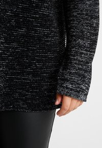 Ruka spočívající podél těla, oblečená v černobílé texturované dlouhé tričko a hladké černé kalhoty na jednobarevném pozadí.