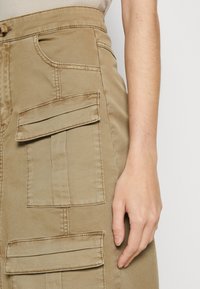 Close-up van een hand van een persoon die rust naast een tan rok met meerdere gestikte zakflappen en een knoopsluiting in de taille.