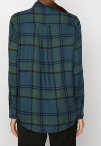 Osoba nosząca koszulę w niebiesko-zieloną kratę z długim rękawem, pokazana od tyłu, w ciemnych spodniach na jednolitym jasnym tle.