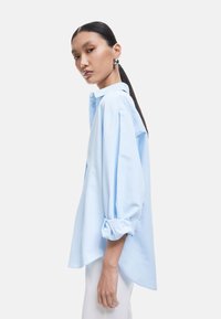 Camicia button-up azzurro chiaro a maniche lunghe con una vestibilità rilassata, caratterizzata da polsini arrotolati e orlo curvo per una silhouette casual.