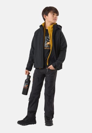 Schwarze Softshell-Jacke mit gelber Innenfütterung, schwarzes T-Shirt mit Grafik, schwarze Cargo-Hose und schwarze Wanderschuhe. Hält eine schwarze Wasserflasche.