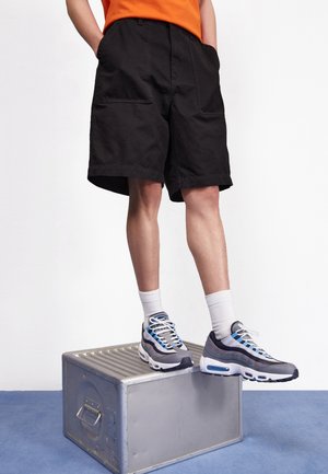 Person iført sorte shorts, hvide crew-sokker og gråblå sneakers, stående med den ene fod på en metalboks mod en hvid baggrund.